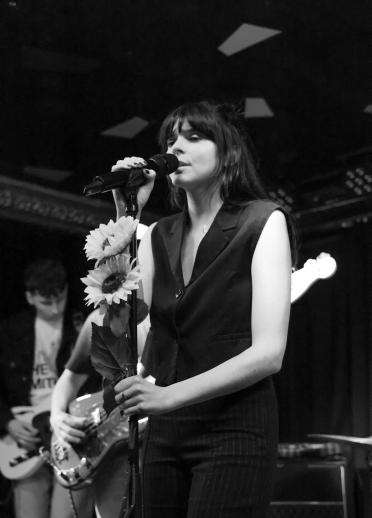Black and white image of young female with dark air and fringe sings into a microphone which is decorated with sunflowers