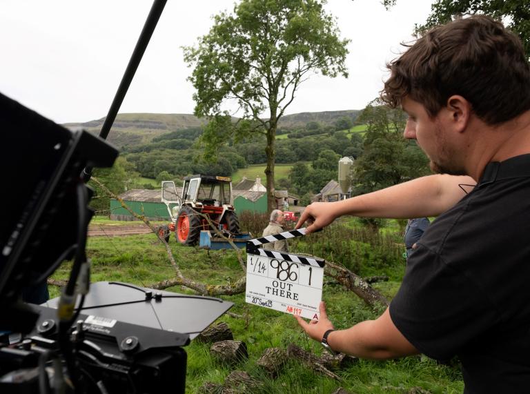 young man raises a clapper board in front of a camera with a tractor and farmland in the background