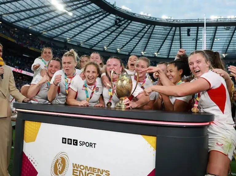 Rugby team celebrating with a trophy on stage at a stadium with a presenter holding a microphone nearby