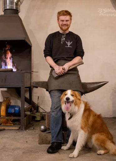 Blacksmith smiling with a hammer in hand, standing by an anvil and forge, alongside a happy, seated dog