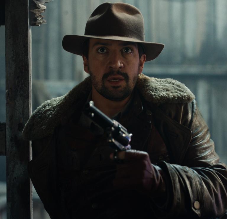 Close up still of Lee Scoresby (Lin-Manuel Miranda) from His Dark Materials (Series 2) (Bad Wolf). Lin-Manuel is wearing wearing a brown hat, brown jacket and is holding a metal handgun.