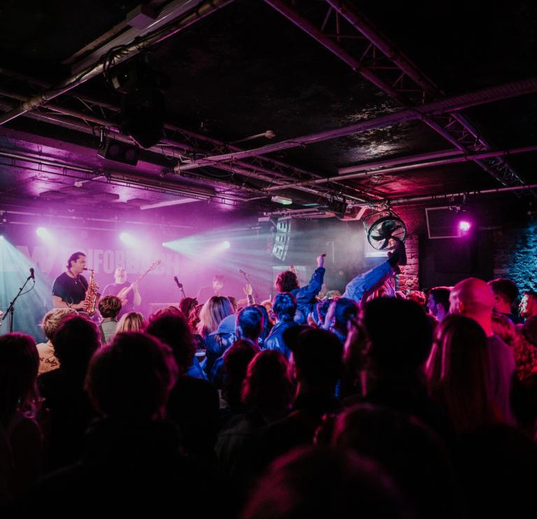 Person crowdsurfing in a dark room lit up by stage lights while a band plays on stage at Club Ifor Bach for the BBC 6 Music Festival