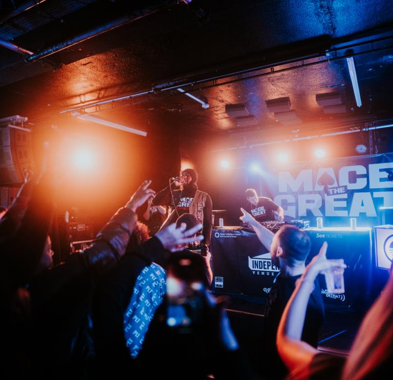 Photo of a dark gig venue. In the foreground, you see the audiences' hands in the air. On stage, Mace the Great performs under bright orange and blue lights. 