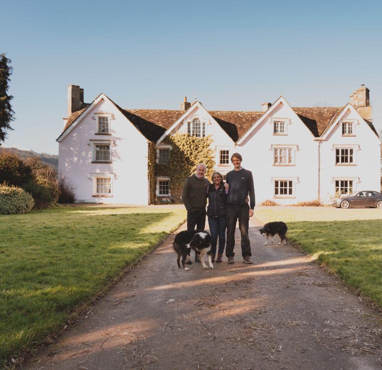 A man standing with a woman and a younger man in front of a large pink farm house on a sunny day