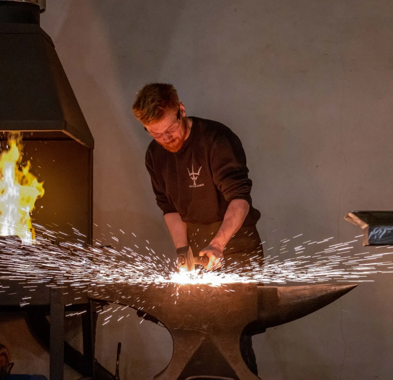 Blacksmith forging metal on an anvil with sparks flying and a furnace ablaze in the background