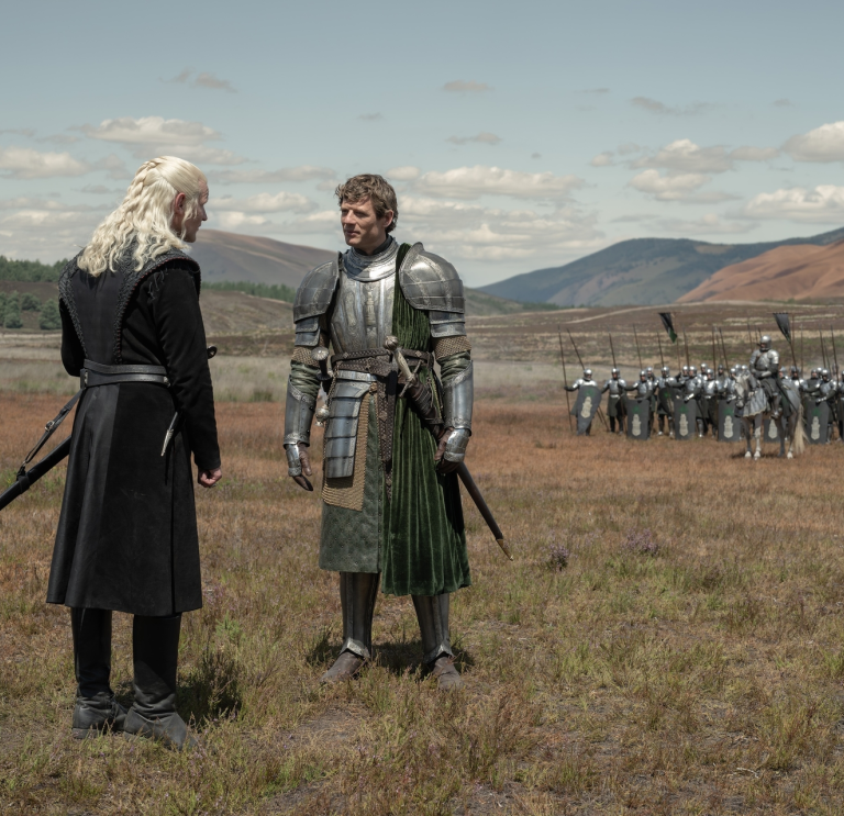 Two armored figures facing each other on a grassy field with soldiers and hills in the background