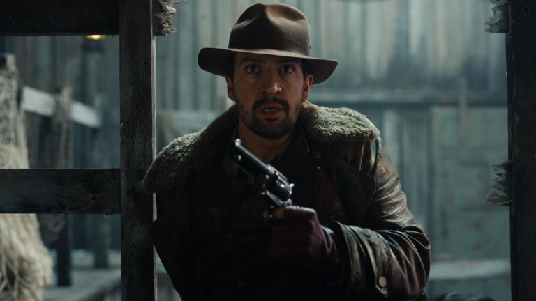 Close up still of Lee Scoresby (Lin-Manuel Miranda) from His Dark Materials (Series 2) (Bad Wolf). Lin-Manuel is wearing wearing a brown hat, brown jacket and is holding a metal handgun.