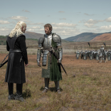 Two armored figures facing each other on a grassy field with soldiers and hills in the background
